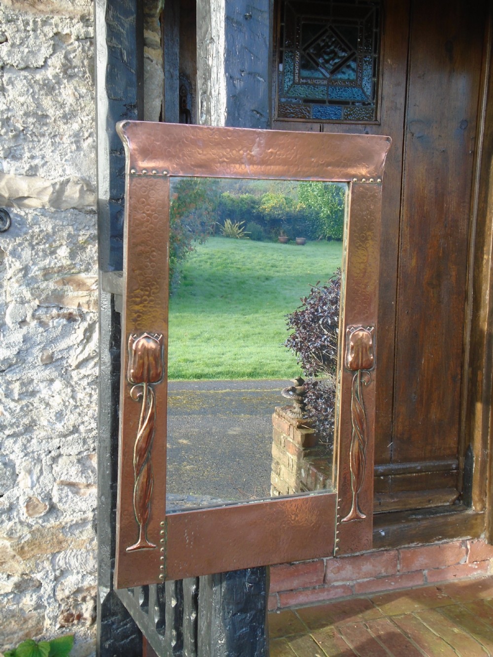 A rectangular Arts and Crafts wall mirror featuring a beaten copper frame with riveted corners and embossed tulip motifs along the side panels.