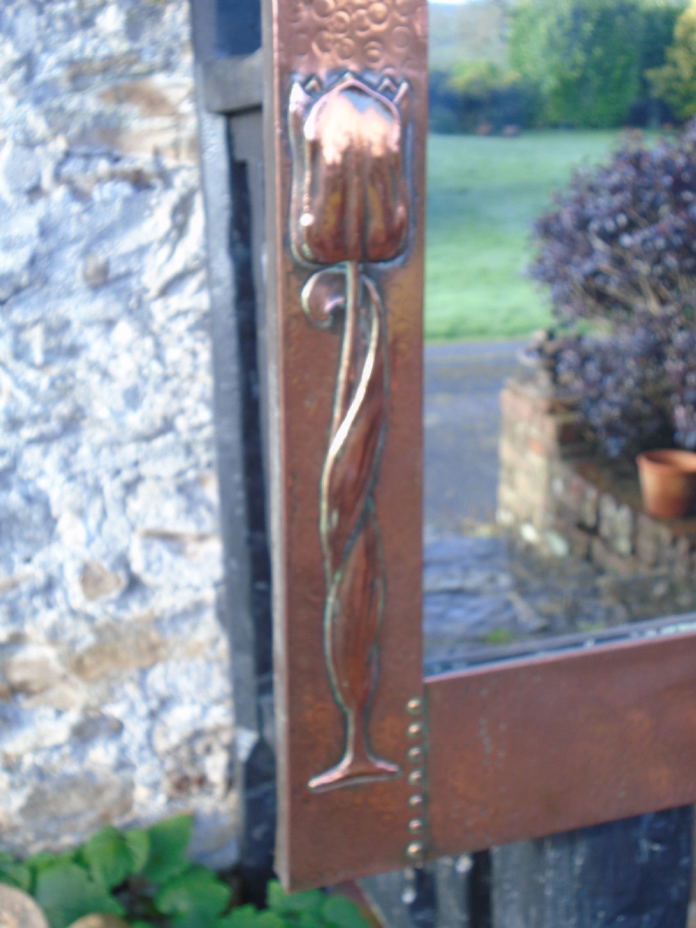 A rectangular Arts and Crafts wall mirror featuring a beaten copper frame with riveted corners and embossed tulip motifs along the side panels. View VI