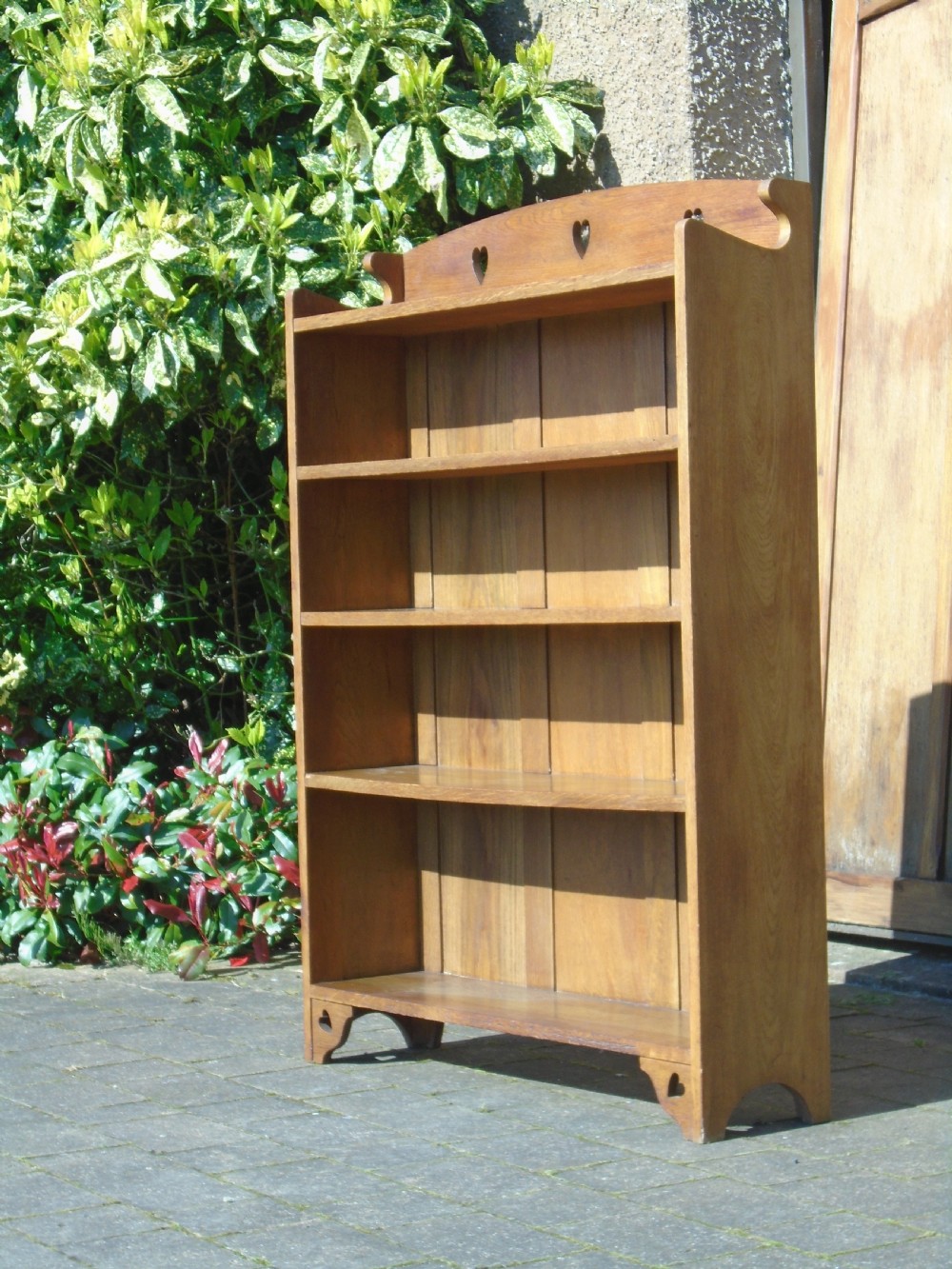 A freestanding Arts and Crafts medium oak open bookcase featuring four shelves, a shaped top gallery with cut-out heart motifs, and cut-out heart details on the base sides.