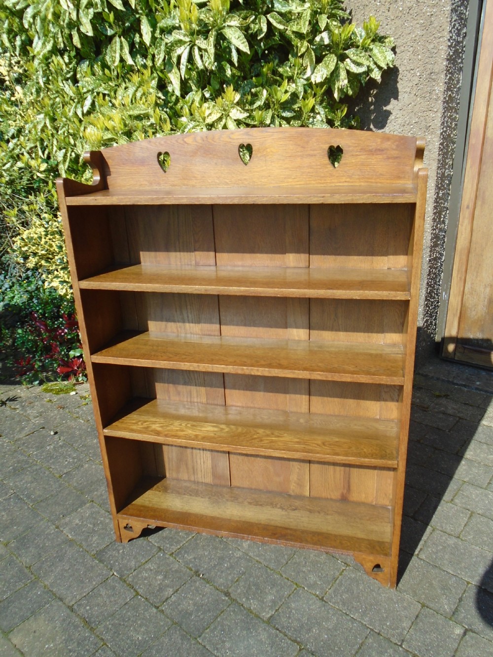 A freestanding Arts and Crafts medium oak open bookcase featuring four shelves, a shaped top gallery with cut-out heart motifs, and cut-out heart details on the base sides. View II