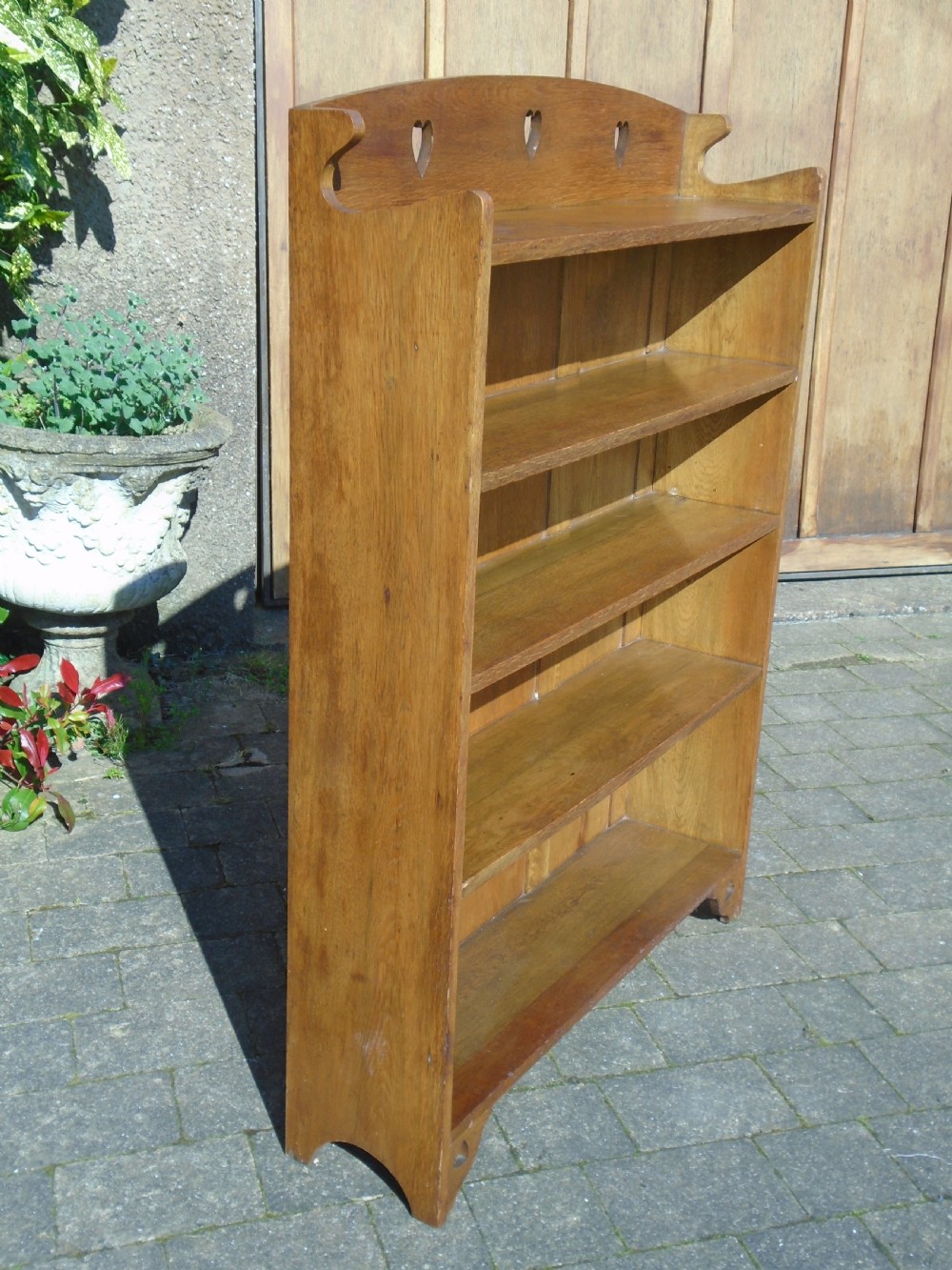 A freestanding Arts and Crafts medium oak open bookcase featuring four shelves, a shaped top gallery with cut-out heart motifs, and cut-out heart details on the base sides. View III