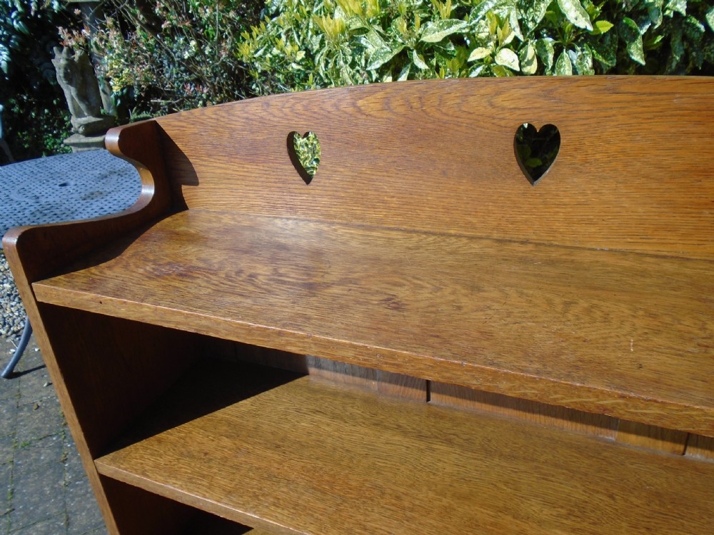 A freestanding Arts and Crafts medium oak open bookcase featuring four shelves, a shaped top gallery with cut-out heart motifs, and cut-out heart details on the base sides. View IV