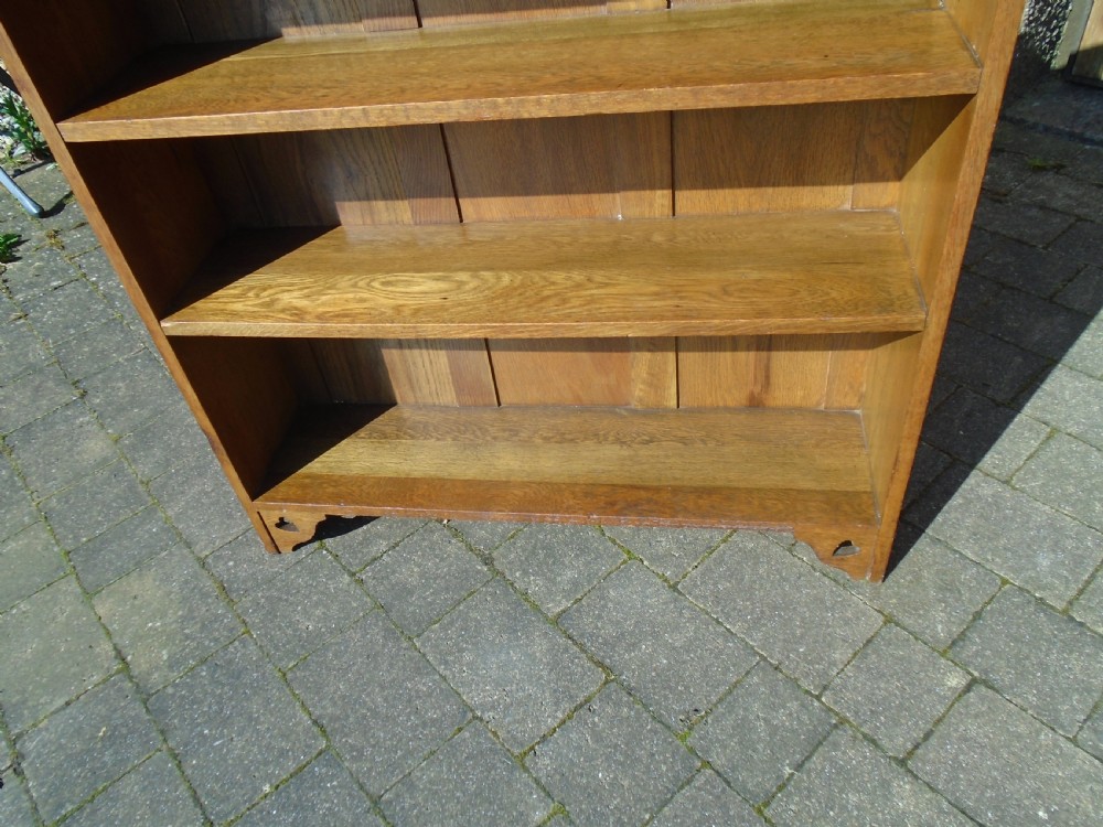 A freestanding Arts and Crafts medium oak open bookcase featuring four shelves, a shaped top gallery with cut-out heart motifs, and cut-out heart details on the base sides. View VI