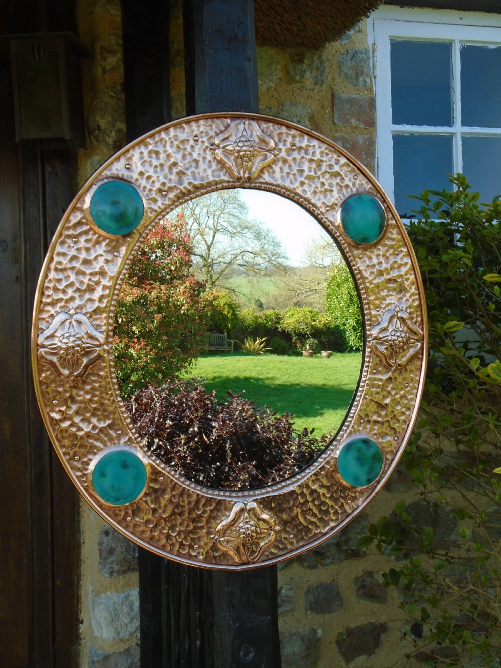 An oval Arts and Crafts wall mirror featuring a hand-hammered copper frame decorated with repousse floral motifs and four turquoise round cabochons.