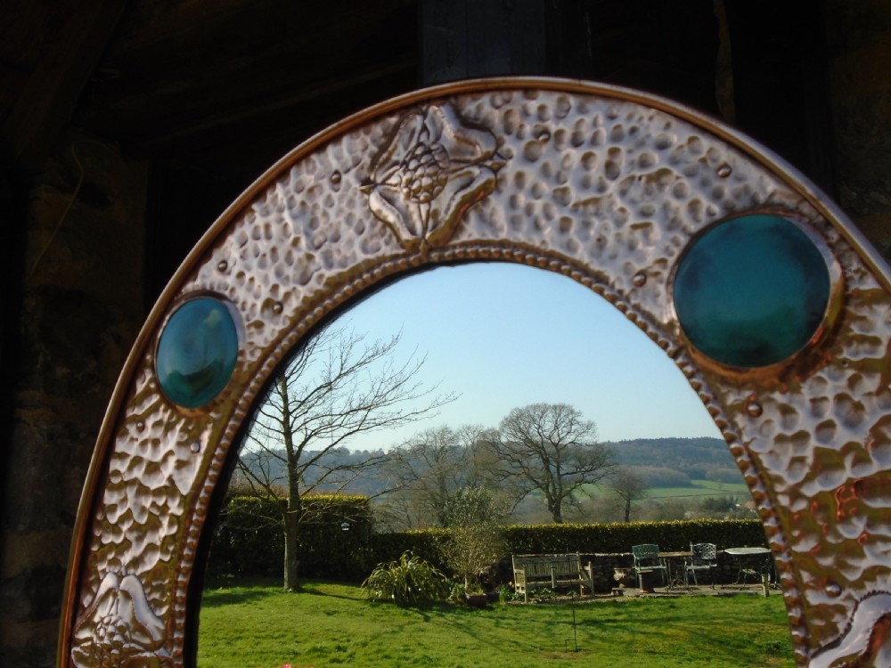 An oval Arts and Crafts wall mirror featuring a hand-hammered copper frame decorated with repousse floral motifs and four turquoise round cabochons. View X