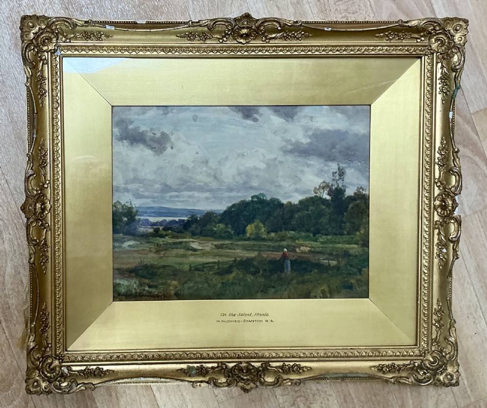 An antique framed watercolour painting by Sir Herbert Hughes-Stanton showing a rural Hampshire landscape under a cloudy sky in a carved gold gilt frame.