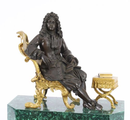 fig 3. A nineteenth-century French mantel clock featuring a green malachite base with gilt bronze mounts, a white enamel dial with Roman numerals, and a dark bronze figure seated on a gilt chair.