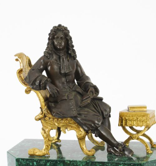 fig 4. A nineteenth-century French mantel clock featuring a green malachite base with gilt bronze mounts, a white enamel dial with Roman numerals, and a dark bronze figure seated on a gilt chair.