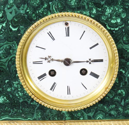 fig 5. A nineteenth-century French mantel clock featuring a green malachite base with gilt bronze mounts, a white enamel dial with Roman numerals, and a dark bronze figure seated on a gilt chair.