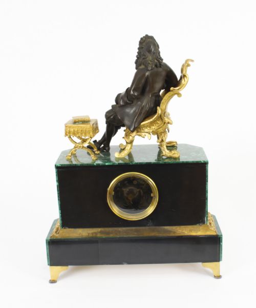 fig 6. A nineteenth-century French mantel clock featuring a green malachite base with gilt bronze mounts, a white enamel dial with Roman numerals, and a dark bronze figure seated on a gilt chair.