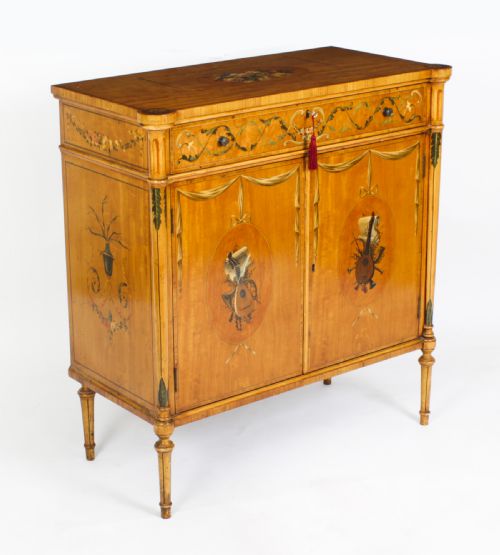 fig 2. A pair of 19th-century Adam Revival painted satinwood side cabinets featuring painted oval musical medallions, draped garlands, single top drawers, double cupboards, and turned tapered legs.