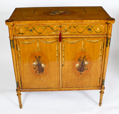 fig 3. A pair of 19th-century Adam Revival painted satinwood side cabinets featuring painted oval musical medallions, draped garlands, single top drawers, double cupboards, and turned tapered legs.