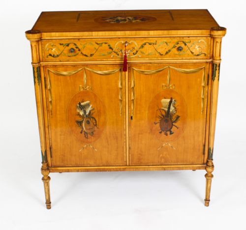 fig 4. A pair of 19th-century Adam Revival painted satinwood side cabinets featuring painted oval musical medallions, draped garlands, single top drawers, double cupboards, and turned tapered legs.