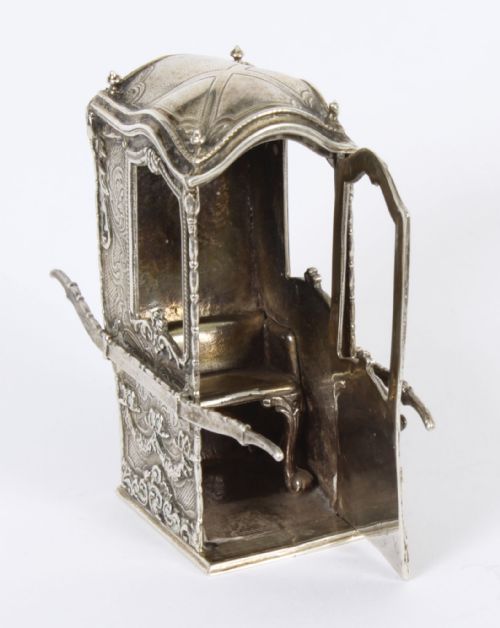 fig 1. A detailed 19th-century silver miniature sedan chair featuring ornate Rococo floral chasing, side carrying poles, a shaped roof, and window openings.