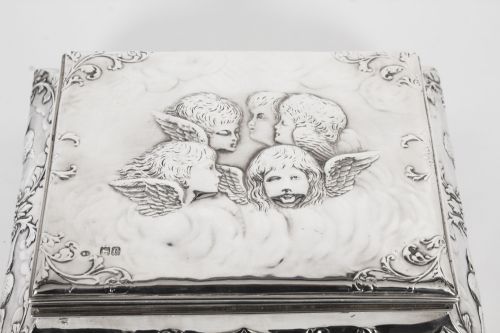 fig 2. A rectangular sterling silver casket featuring relief repousse cherub heads on the hinged lid and scrollwork along the flared base.