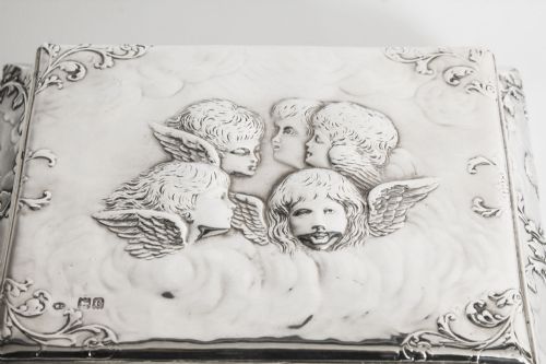 fig 4. A rectangular sterling silver casket featuring relief repousse cherub heads on the hinged lid and scrollwork along the flared base.