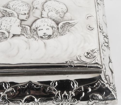 fig 6. A rectangular sterling silver casket featuring relief repousse cherub heads on the hinged lid and scrollwork along the flared base.