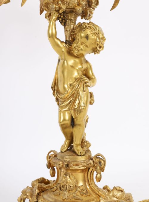 fig 3. A French gilt bronze table lamp featuring a sculpted cherub holding lily-shaped light sockets on a Rococo scrolled base.