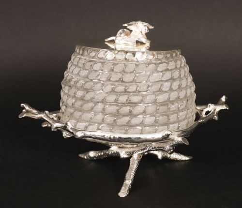 fig 1. A glass beehive-shaped honey jar with a silver-plated lid featuring a bee finial, resting on a silver-plated stand formed as intertwined tree branches.