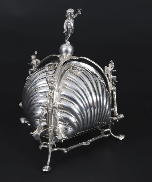 fig 1. An antique silver-plated folding biscuit box featuring scalloped shell compartments, a top cherub finial, and decorative figurative side supports.