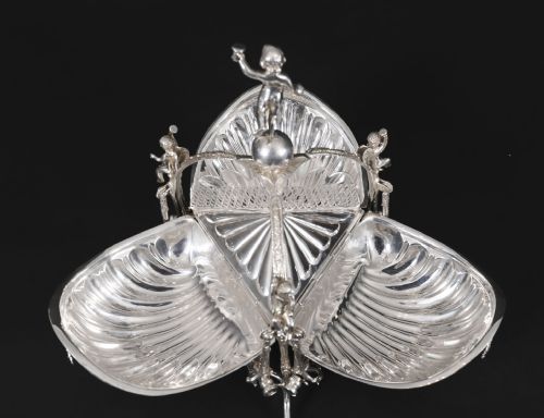 fig 3. An antique silver-plated folding biscuit box featuring scalloped shell compartments, a top cherub finial, and decorative figurative side supports.