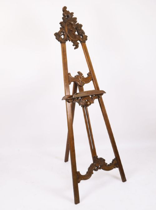 fig 1. An antique carved walnut artist easel featuring an ornate Rococo cresting, an adjustable shelf, and scrolled leg supports.