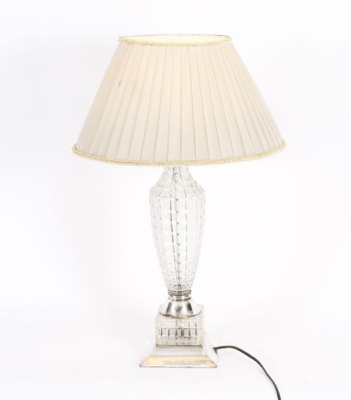 fig 1. An Art Deco table lamp featuring a faceted clear cut crystal stem, a square metal pedestal base, and a cream pleated fabric lampshade.