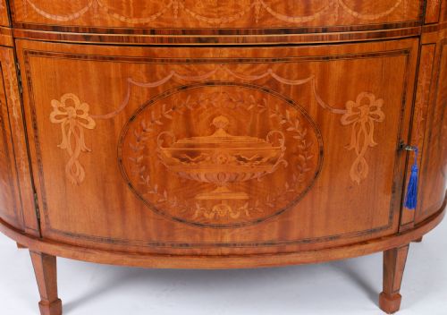 fig 2. A semicircular antique satinwood demilune cabinet featuring urn and swag marquetry inlays, a top drawer with a blue tassel key, and slender tapered legs.