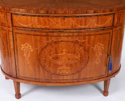 fig 3. A semicircular antique satinwood demilune cabinet featuring urn and swag marquetry inlays, a top drawer with a blue tassel key, and slender tapered legs.