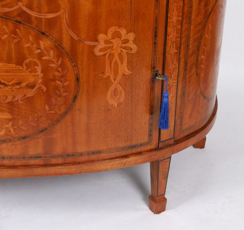 fig 4. A semicircular antique satinwood demilune cabinet featuring urn and swag marquetry inlays, a top drawer with a blue tassel key, and slender tapered legs.
