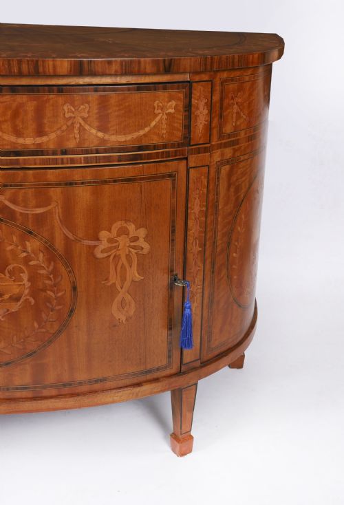 fig 7. A semicircular antique satinwood demilune cabinet featuring urn and swag marquetry inlays, a top drawer with a blue tassel key, and slender tapered legs.