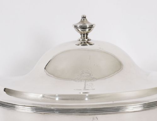fig 4. A George III sterling silver tureen with two looped handles, a domed lid, a pedestal base, and an engraved heraldic crest.