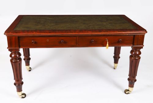 fig 1. A William IV mahogany writing desk featuring an inset green leather surface, two frieze drawers with knob handles, turned fluted legs, and brass castors.