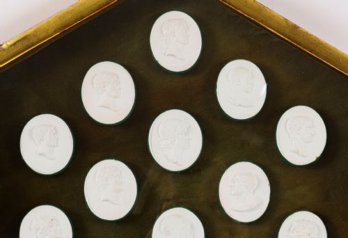 fig 1. A hexagonal giltwood frame housing thirteen classical white plaster profile intaglios mounted against a dark green background.