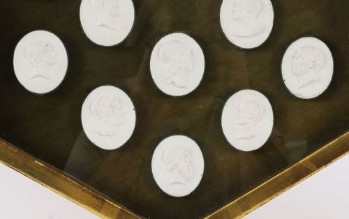 fig 2. A hexagonal giltwood frame housing thirteen classical white plaster profile intaglios mounted against a dark green background.