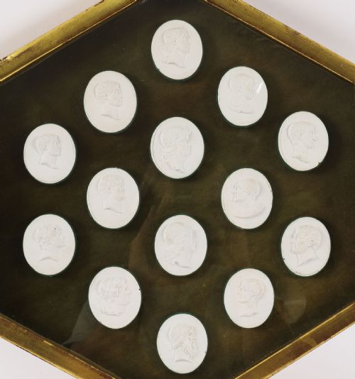 fig 3. A hexagonal giltwood frame housing thirteen classical white plaster profile intaglios mounted against a dark green background.