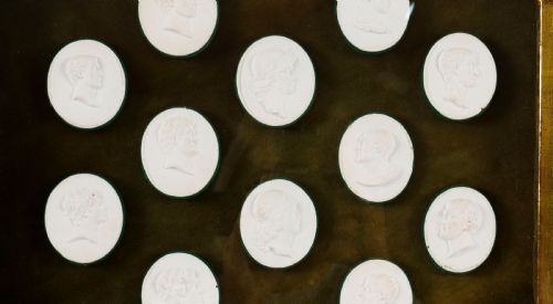 fig 4. A hexagonal giltwood frame housing thirteen classical white plaster profile intaglios mounted against a dark green background.