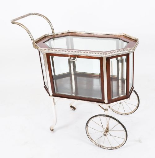 fig 1. An Art Deco drinks trolley featuring an octagonal glass top with metal gallery trim, wooden side frames, spoked wheels, and a drop-down door.