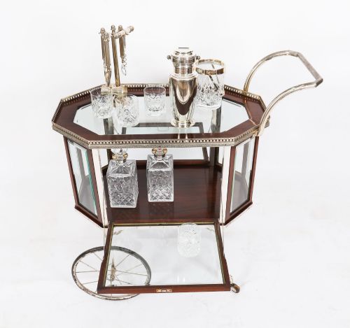 fig 3. An Art Deco drinks trolley featuring an octagonal glass top with metal gallery trim, wooden side frames, spoked wheels, and a drop-down door.