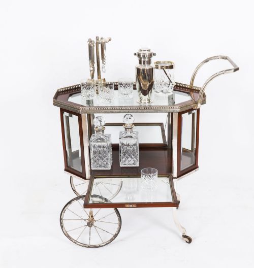 fig 5. An Art Deco drinks trolley featuring an octagonal glass top with metal gallery trim, wooden side frames, spoked wheels, and a drop-down door.