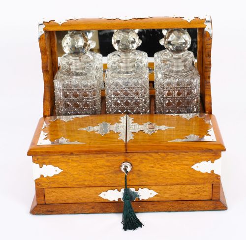 fig 1. An antique blonde oak tantalus featuring three square cut-glass decanters set against a mirror back, decorative silver-plated corner mounts, and a lower drawer with a key.