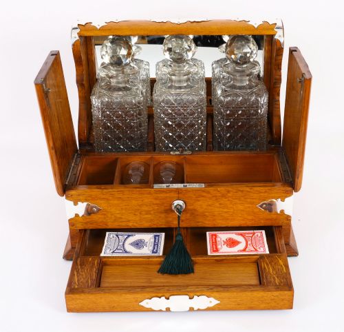 fig 7. An antique blonde oak tantalus featuring three square cut-glass decanters set against a mirror back, decorative silver-plated corner mounts, and a lower drawer with a key.