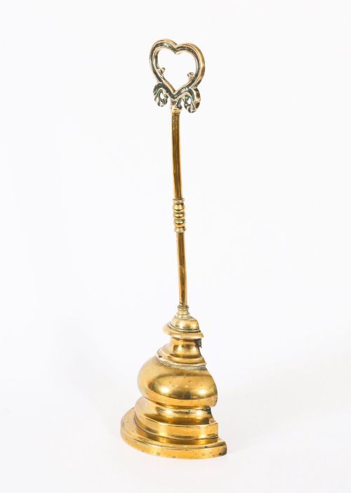 fig 1. A 19th-century Victorian brass door stop featuring a tall shaft topped with a heart-shaped handle and supported by a heavy, tiered circular base.