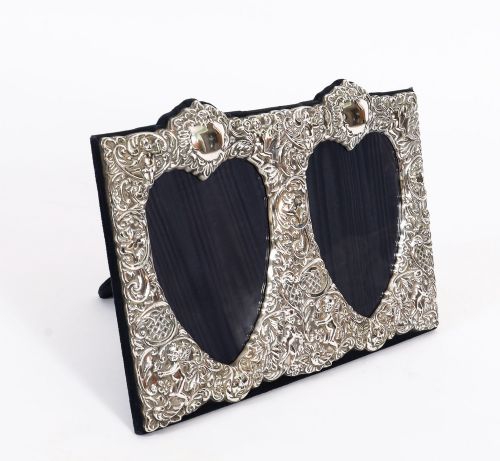 fig 1. An antique sterling silver double aperture photo frame featuring heart-shaped openings, intricate repousse floral scrollwork, and cherub details mounted on dark velvet.