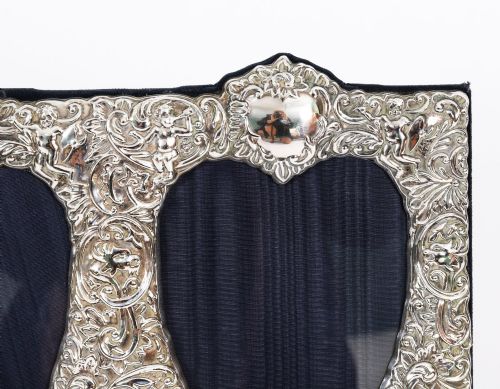 fig 2. An antique sterling silver double aperture photo frame featuring heart-shaped openings, intricate repousse floral scrollwork, and cherub details mounted on dark velvet.