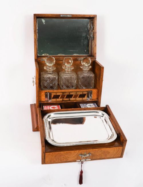 fig 1. A 19th-century Victorian figured oak three-bottle tantalus open to reveal three cut glass decanters, a fitted cribbage board, and a drop-front drawer containing a silver-plated tray and playing cards.