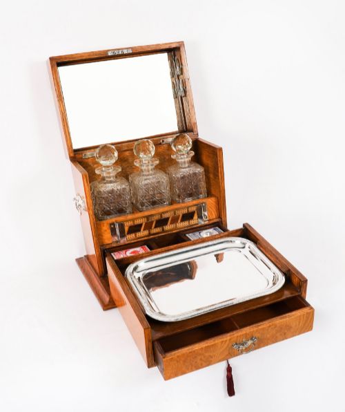 fig 2. A 19th-century Victorian figured oak three-bottle tantalus open to reveal three cut glass decanters, a fitted cribbage board, and a drop-front drawer containing a silver-plated tray and playing cards.