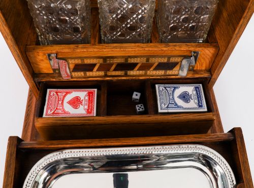 fig 5. A 19th-century Victorian figured oak three-bottle tantalus open to reveal three cut glass decanters, a fitted cribbage board, and a drop-front drawer containing a silver-plated tray and playing cards.