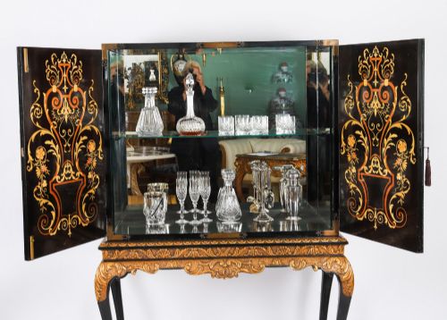 fig 3. A black lacquered and inlaid wooden cocktail cabinet featuring ornate floral marquetry and decorative brass straps, resting on a carved gilded stand with cabriole legs.