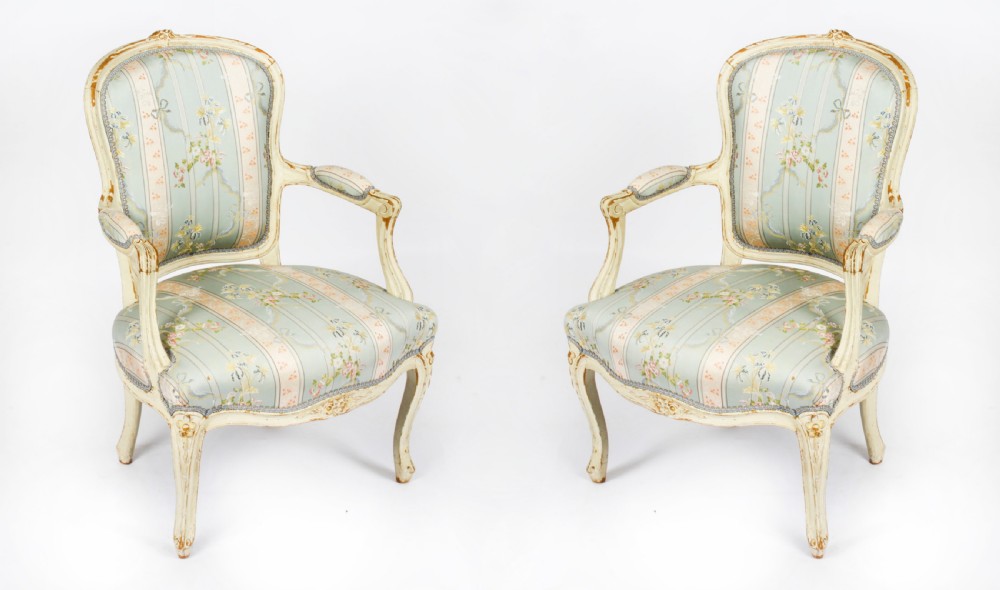 A pair of 19th-century French Louis Revival armchairs featuring distressed cream-painted carved wooden frames and striped pastel floral silk upholstery.